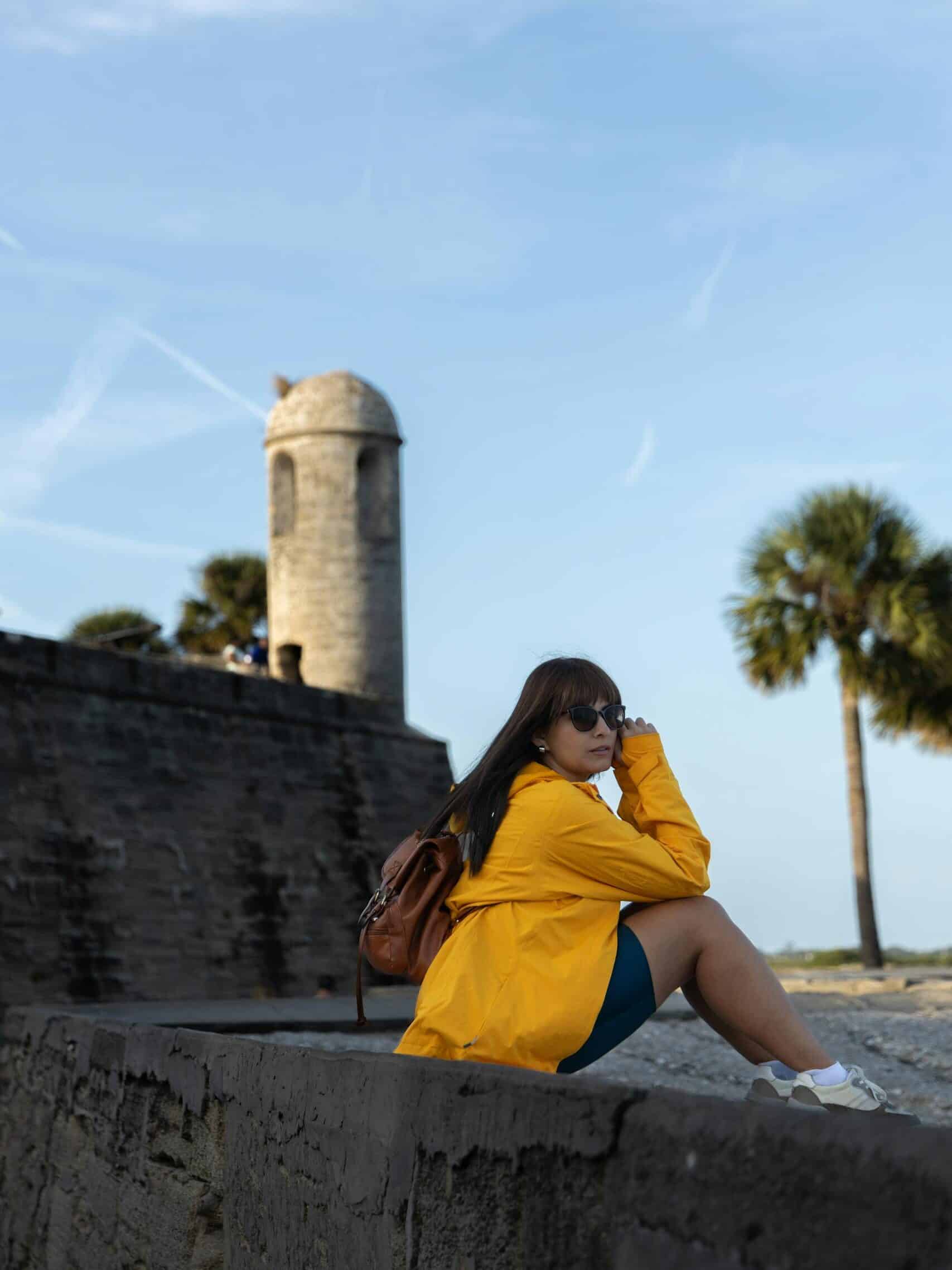 A woman in a yellow jacket sits by a historic tower under a clear blue sky.