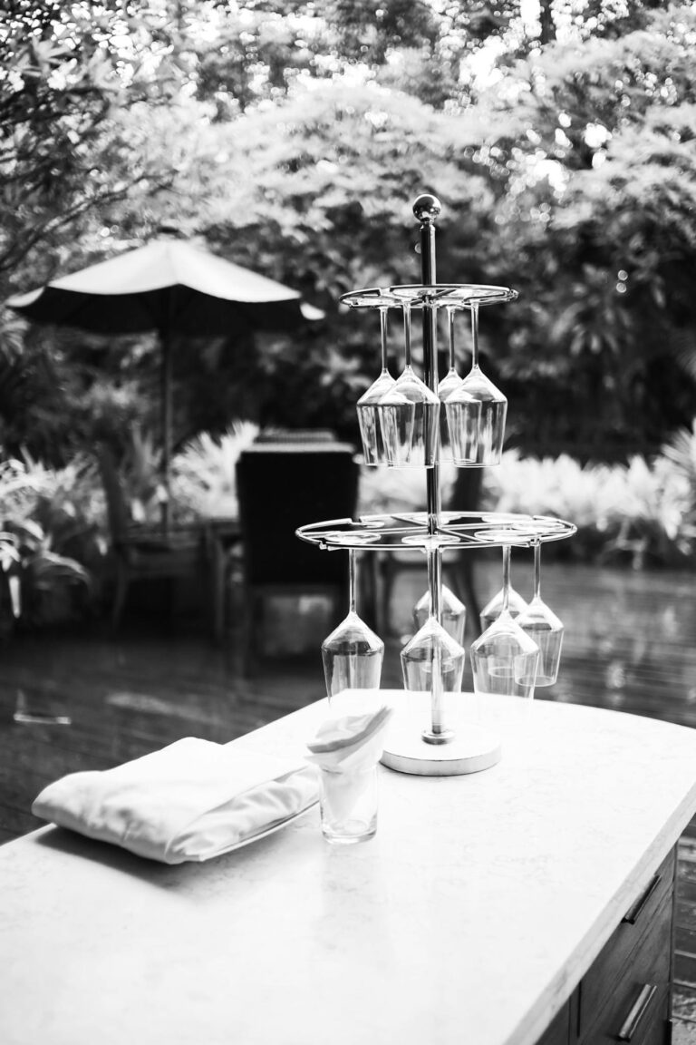 Black and white image of an outdoor bar setting with stacked wine glasses and lush background.